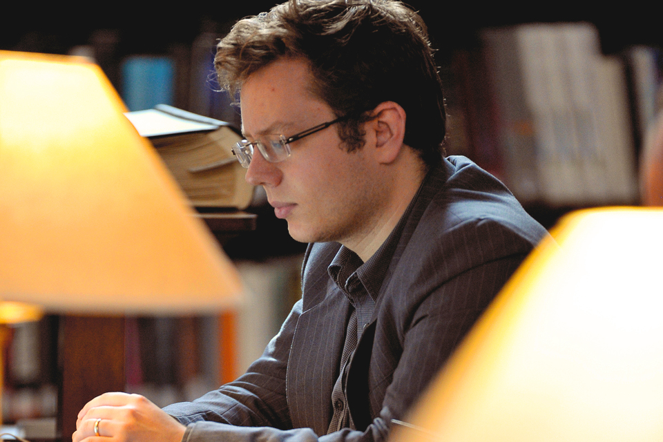 A man with dark hair and glasses, studying in a library with small lamps in the foreground.